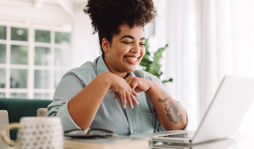 A woman sits a table indoors and smiles while looking at an open laptop.