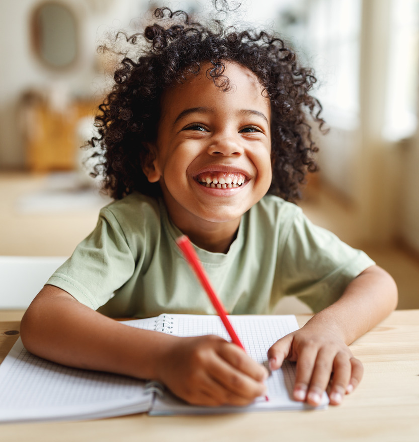 A small boy with curly hair looks up and smiles. He holds a pencil about to write in a notebook.