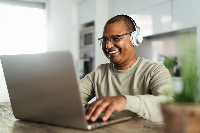 A man with white headphones sits in front of an open laptop smiling. He is indoors sitting at a table.