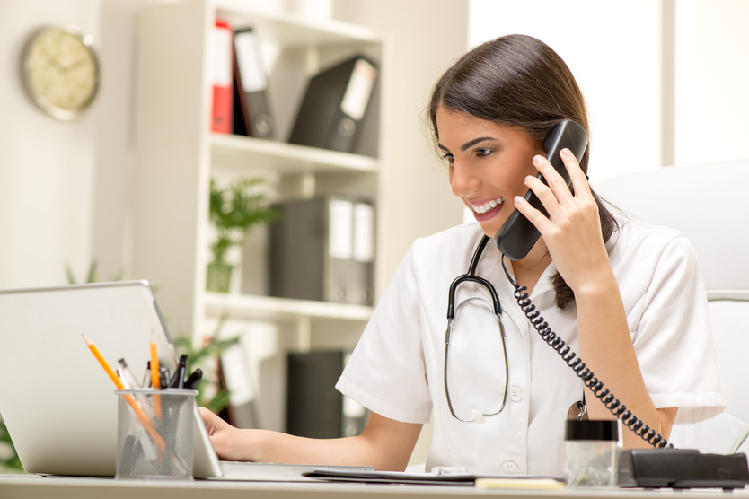 A doctor talks on a corded telephone. She is smiling and has a stethoscope around her neck.