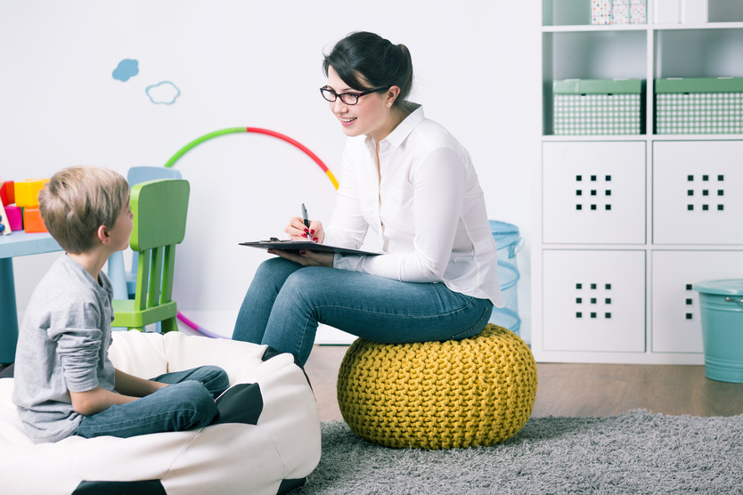 A woman with a clipboard sits on a cushion in a play room, talking with a young boy.