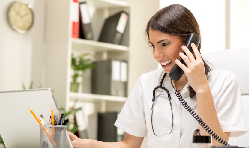 A doctor talks on a corded telephone. She is smiling and has a stethoscope around her neck.