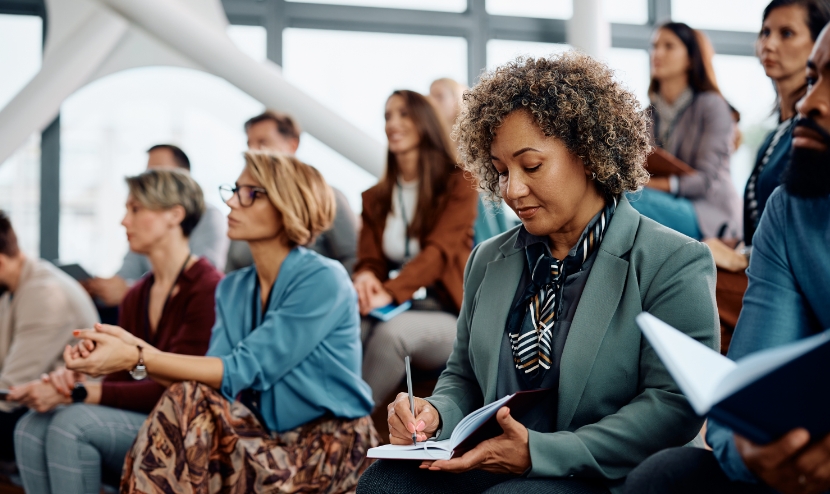 Several people sit in an auditorium watching a presentation. A woman in the foreground with a light blue jacket takes notes in a notebook.