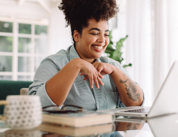 A woman sits a table indoors and smiles while looking at an open laptop.