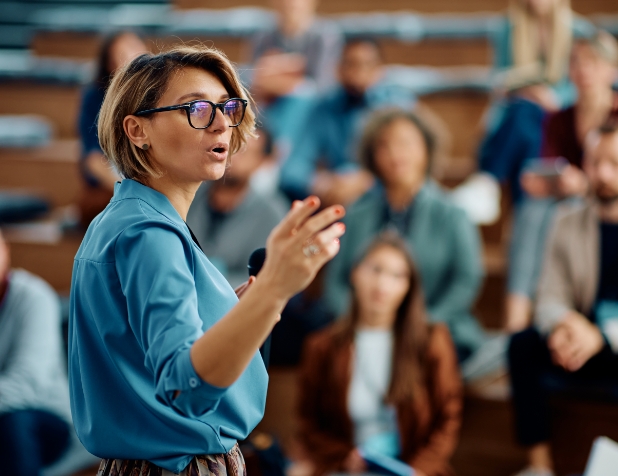 A woman in a blue shirt and black glasses stands in front of an audience. She is gesturing with her hand while talking.