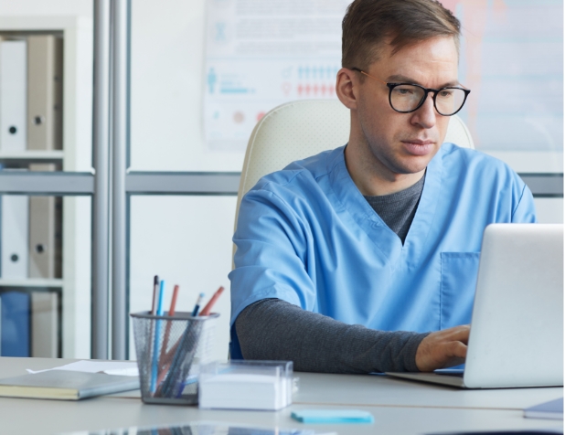 A doctor in blue scrubs sits behind a table at a laptop, concentrating on his work.