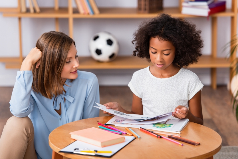 A young child shares her drawing work with a woman. They are both at a low table with paper and colored pencils.