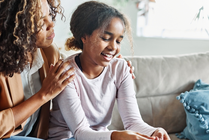 A mother and daughter sit on a couch talking. The mother has her hands on the girl's shoulders while she listens.