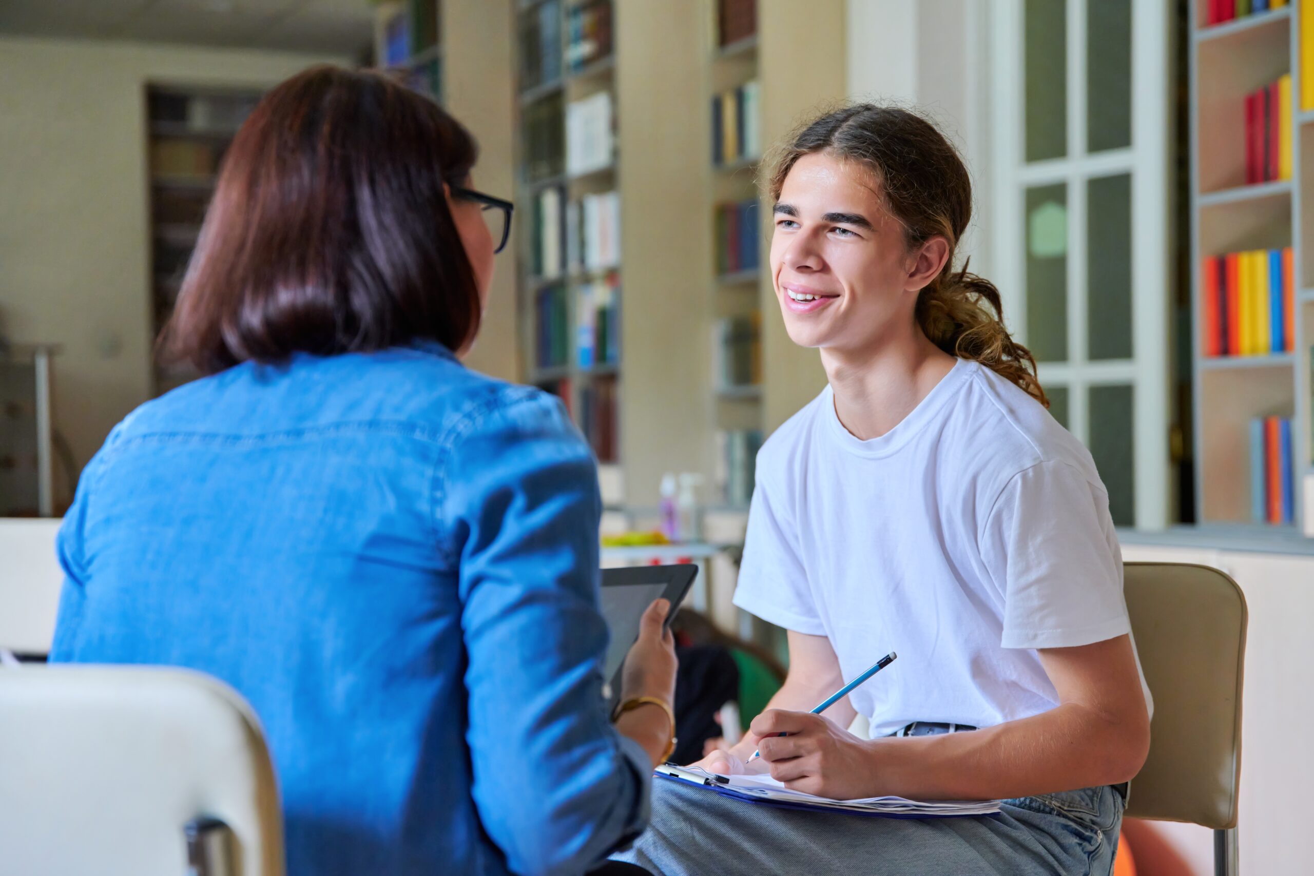 Woman school psychologist teacher talking and helping student, male teenager, inside the library. Mental health of adolescents, psychology, social issues, professional help of counselor